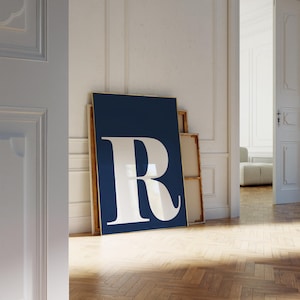 May include: A framed navy blue art print featuring a large white letter "R". The artwork is leaning against a white wall, with a glimpse of a room in the background. The print has a gold frame.