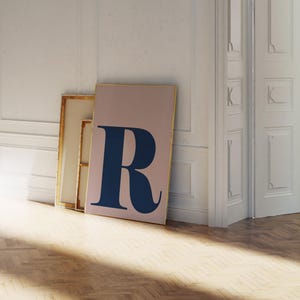 以下が含まれることがあります： ブラッシュピンクの背景に、濃いネイビーブルーの文字「R」が描かれたフレーム入りアートプリント。白い壁に立てかけられており、他の額装された作品の隣にあります。フレームは金色の仕上げです。部屋には寄木張りの床があります。