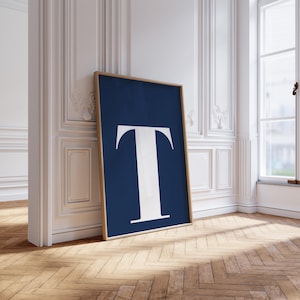 May include: A framed art print featuring a large white letter "T" against a navy blue background. The artwork is displayed in a room with white walls and a wooden floor, leaning against the wall.