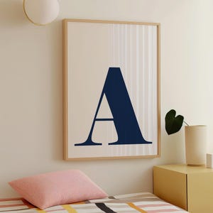 May include: A framed print featuring a large, navy blue letter "A" against a cream-colored background. The artwork is encased in a light wood frame. The print is displayed in a room with a bed and a side table.