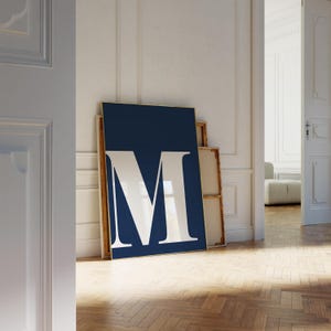 May include: A framed navy blue art print with a large white letter "M". The artwork is propped against a white wall, with other framed pieces visible. The room has a wooden floor and an open doorway.