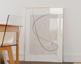 May include: Abstract art print in a gold frame. The artwork features black lines on a beige background. The print is displayed against a white wall, with a wooden chair and floor visible in the foreground.