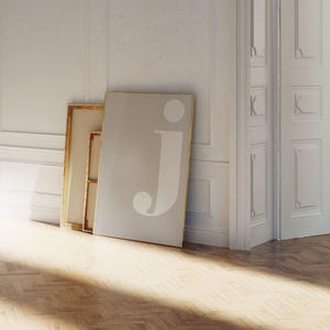 May include: A large, rectangular framed print with the letter "j" in white against a light gray background. The print leans against a white wall, next to a framed canvas. The floor is made of light-colored wood.