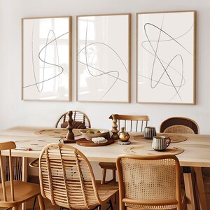 May include: Three framed abstract art prints with thin black lines on a cream background. The prints are displayed above a wooden dining table with woven chairs and decorative items.