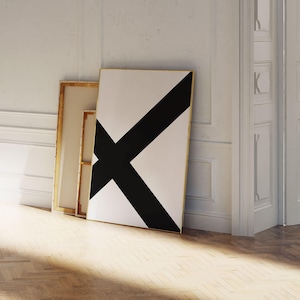 May include: Three framed artworks lean against a white wall. The largest features a bold black X on a white background, framed in gold. The other two have a gold frame and a canvas. The floor is light-colored wood.