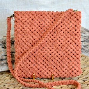 May include: A square-shaped crossbody bag crafted from terracotta-colored cord in a dense macrame knot pattern. The bag features a matching braided shoulder strap attached with metallic swivel clasps at the top corners and a flat, structured base.