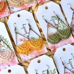 May include: Assortment of handmade crocheted earrings with triangle-shaped metal hoops. The earrings are in various colors, including pink, orange, green, and purple. The earrings are displayed on white cards with the text "River Jewelry" printed on them.