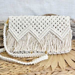 May include: An off-white macrame crossbody bag featuring a complex knotted diamond pattern across the flap, a chevron-style fringe trim along the bottom edge, and a matching braided shoulder strap attached with small gold-toned metal hardware.