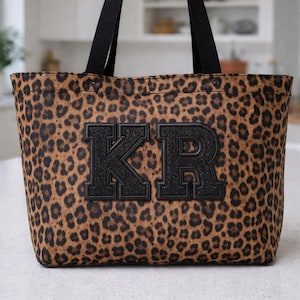 Könnte beinhalten: Eine Tragetasche mit Leopardenmuster und schwarzen Griffen sowie den Buchstaben "KR" in schwarzer Schrift im College-Stil. Die Tasche besteht aus einem braunen und beigefarbenen Stoff mit Flecken und steht auf einer weißen Arbeitsplatte.