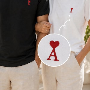 Può includere: Una t-shirt nera con un cuore e la lettera K ricamati in rosso, e una t-shirt bianca con un cuore e la lettera A ricamati in rosso. Un riquadro circolare mostra una vista ingrandita del ricamo del cuore rosso e della lettera A sulla t-shirt.