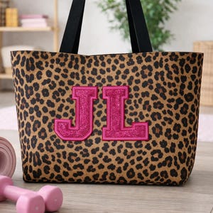 Puede incluir: Un bolso tote con estampado de leopardo, asas negras y un monograma rosa brillante. La bolsa está junto a una botella de agua rosa, una esterilla de yoga enrollada, pesas y una toalla blanca enrollada. Un accesorio elegante.