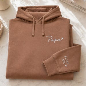 May include: A brown hoodie with a drawstring hood and a kangaroo pocket. The word "Papa" is embroidered on the chest in white script, with a small heart. The sleeve cuff has the names "Elias" and "Aya" embroidered in white, with a heart.