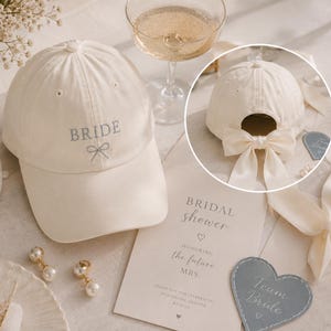 May include: A cream-coloured baseball cap featuring the embroidered word BRIDE and a small bow icon on the front. The back of the cap includes a matching cream-coloured fabric bow tied around the adjustable strap opening, adding a decorative detail.