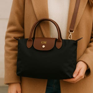 May include: Black handbag with brown leather detailing on the handles and flap. The flap is embossed with the initials "GM". It features a detachable brown shoulder strap with gold-coloured hardware. The bag is held by a person wearing a tan coat.