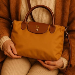 May include: Mustard yellow handbag with brown leather detailing. Includes a top handle and a detachable shoulder strap. The flap closure is embossed with the initials "ER". The bag is made of nylon.