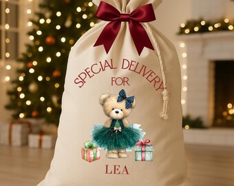 May include: A beige Christmas gift bag with a burgundy bow at the top. The bag features a teddy bear wearing a teal tutu and a blue bow. The words "SPECIAL DELIVERY FOR LEA" are printed on the bag, along with two wrapped gifts.
