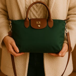 May include: A green handbag with brown leather detailing, including the handles and flap. The bag has a shoulder strap and a gold-coloured clasp. The flap is embossed with the initials "KR".