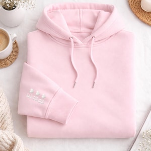 Könnte beinhalten: Ein hellrosa Kapuzenpullover mit Kordelzugkapuze und gerippten Bündchen. Der linke Ärmel hat die gestickten Buchstaben "BBB" mit drei Herzsymbolen. Der Hoodie ist aus weichem Material und wird auf einer weißen Oberfläche präsentiert.