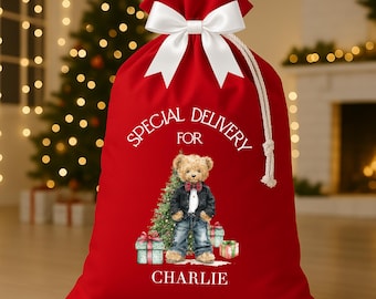 May include: A red Christmas gift bag with a white bow and drawstring closure. The bag features a teddy bear illustration with the text "Special Delivery For" and the name "Charlie". Presents and a Christmas tree are also depicted.