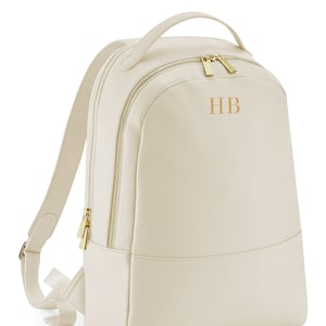 Może przedstawiać: Kremowy plecak ze sztucznej skóry z złotymi zamkami błyskawicznymi i złotym uchwytem. Plecak ma monogram z literami "HB" z przodu.