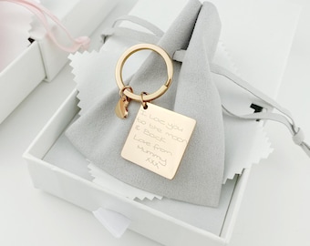 Puede incluir: Un llavero dorado con un dije cuadrado que dice "Te quiero hasta la luna y de regreso Amor de mamá xxx". El llavero está en una pequeña caja blanca con una bolsa de terciopelo gris.