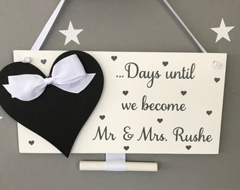 May include: White wooden sign with a black chalkboard heart with a white ribbon bow. The sign says "...Days until we become Mr & Mrs. Rushe" with small hearts around the text.
