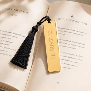 Peut inclure: Un marque-page rectangulaire en métal doré avec le prénom ELIZABETH gravé en lettres capitales. Le marque-page est attaché à un cordon noir avec un pompon en tissu noir assorti, posé sur les pages ouvertes d'un livre imprimé.