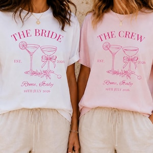 Op de afbeelding: Een wit t-shirt met roze tekst THE BRIDE en een lichtroze t-shirt met roze tekst THE CREW. Beide t-shirts hebben een afbeelding van twee cocktailglazen, de tekst EST. 2026, Rome, Italy, en 19TH JULY 2026 in roze inkt op de voorzijde.