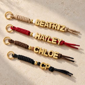 May include: Four keychains featuring gold-toned metal letters spelling BEATRIZ, HAYLEY, CHLOE, and CP. Each keychain has a gold metal ring, a section of braided cord in beige, red, brown, or black, and a single decorative bead before the letters.