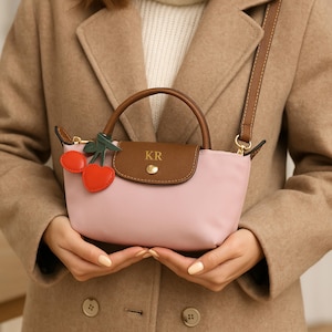 Op de afbeelding: Een roze handtas met een bruin lederen handvat en flap, met de initialen "KR". De tas heeft een afneembare schouderriem en een kersenbedel. De tas wordt vastgehouden door een persoon die een beige jas draagt.