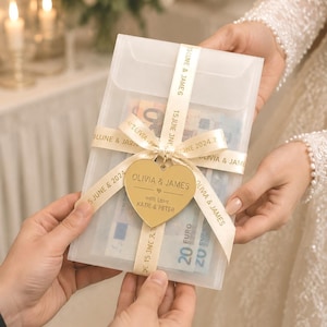 May include: A translucent rectangular envelope containing Euro banknotes, wrapped with a cream-colored ribbon printed with the text OLIVIA & JAMES and 15 JUNE 2024. A gold-colored heart-shaped tag hangs from the ribbon, engraved with OLIVIA & JAMES.