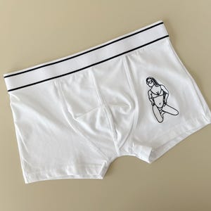 May include: White boxer briefs with a black waistband and a black line drawing of a woman in a bikini on the front.