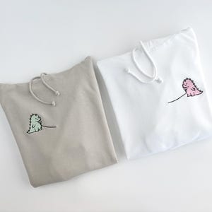 May include: Two hooded sweatshirts with dinosaur embroidery. One sweatshirt is beige with a green dinosaur, the other is white with a pink dinosaur.