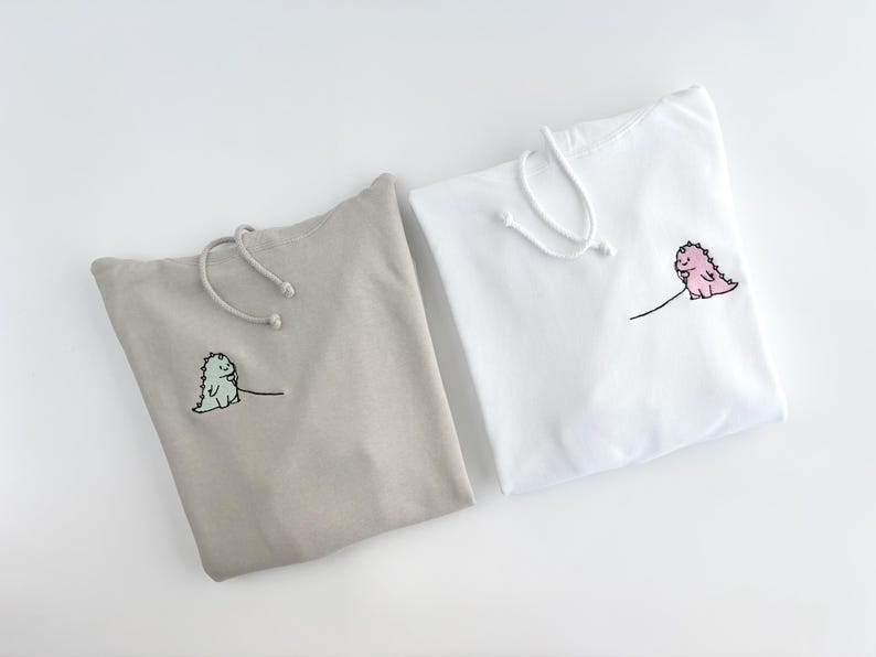 May include: Two hooded sweatshirts with dinosaur embroidery. One sweatshirt is beige with a green dinosaur, the other is white with a pink dinosaur.