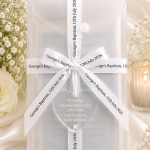May include: A translucent rectangular envelope secured with a white ribbon printed with the text George's Baptism, 12th July 2026. A clear heart-shaped acrylic ornament hangs from the ribbon, inscribed with To George, on your baptism day 12/07/2026.