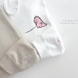 May include: Two hooded sweatshirts, one beige and one white, with embroidered pink dinosaurs and a letter on the sleeves. The white sweatshirt has the text "CUSTOMISED WITH A LETTER OF YOUR CHOICE".