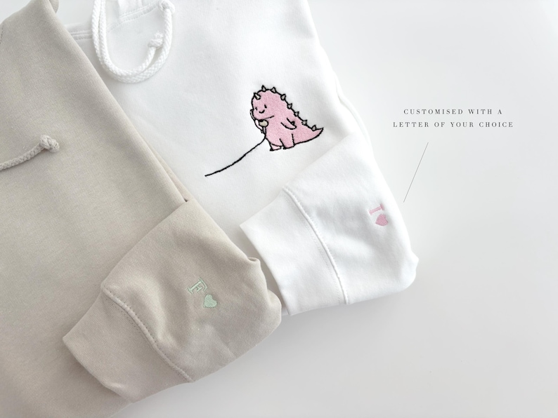 May include: Two hooded sweatshirts, one beige and one white, with embroidered pink dinosaurs and a letter on the sleeves. The white sweatshirt has the text "CUSTOMISED WITH A LETTER OF YOUR CHOICE".