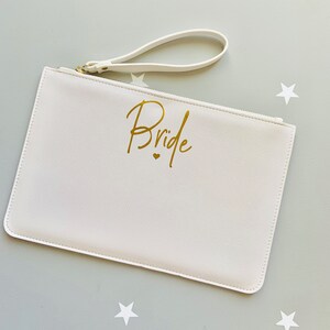 Peut inclure: Pochette blanche avec une fermeture &eacute;clair dor&eacute;e et une inscription dor&eacute;e qui dit "Bride" avec un petit c&oelig;ur.