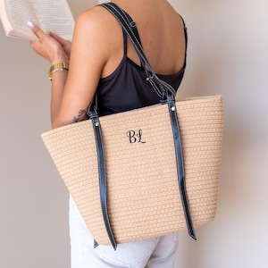 May include: A beige woven tote bag with black straps and the initials "BL" embroidered on the front. The bag is rectangular and has a structured shape. The straps are attached with silver-coloured rivets. The bag is being held.