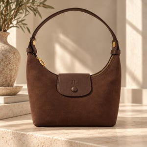 Pode incluir: Uma bolsa hobo de camurça castanho-escura com uma alça arredondada única, fecho de correr em tom dourado e uma aba central com as letras JL gravadas. A bolsa tem uma base estruturada e ferragens em tom dourado nos pontos de fixação da alça.