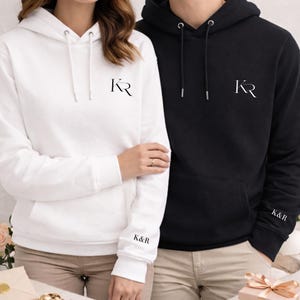 Może przedstawiać: Białe i czarne bluzy z kapturem z pasującymi inicjałami. Biała bluza ma inicjały "KR" na piersi i "K&R" na rękawie. Czarna bluza ma również inicjały "KR" na piersi i "K&R" na rękawie. Obie bluzy mają sznurki.