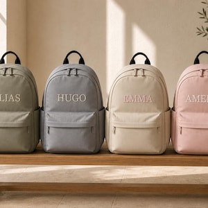 Puede incluir: Cuatro mochilas en verde salvia, gris, beige y rosa claro, cada una con asa superior, bolsillo frontal con cremallera y bolsillos laterales. Los nombres ELIAS, HUGO, EMMA y AMELIA están bordados en letras mayúsculas blancas en el frente.