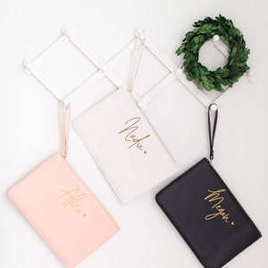 May include: Three personalized cosmetic bags with gold lettering. The bags are white, pink, and black. The names on the bags are Nadia, Allie, and Megan.