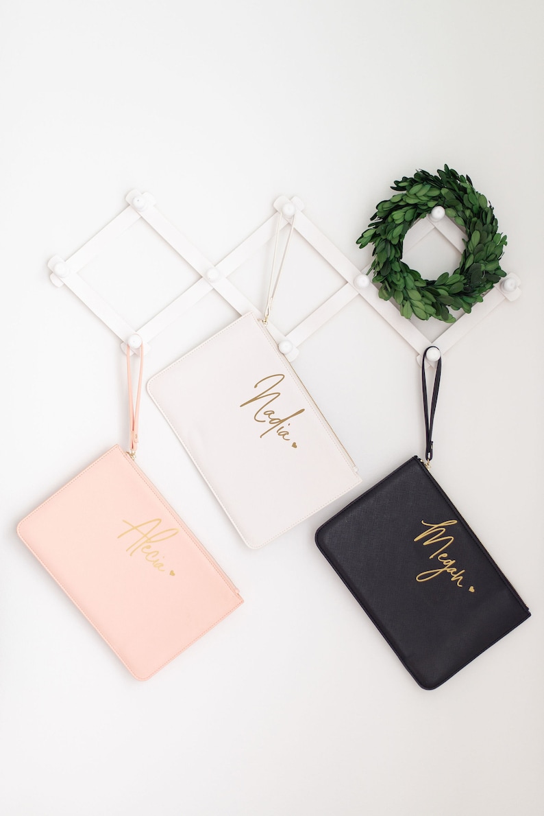 May include: Three personalized cosmetic bags with gold lettering. The bags are white, pink, and black. The names on the bags are Nadia, Allie, and Megan.