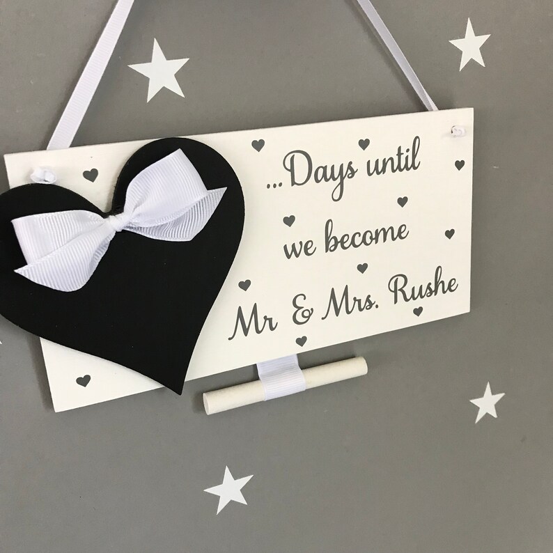 May include: White wooden sign with a black chalkboard heart and a white ribbon bow. The sign says "... Days until we become Mr & Mrs. Rushe" with small hearts around the text.