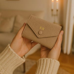 Puede incluir: Una cartera de cuero color beige con cierre de solapa y botón. Las iniciales "KR" están grabadas en dorado en la parte delantera. La cartera está sujeta en las manos de una persona.