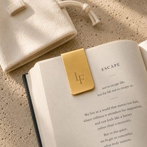 Könnte beinhalten: Ein magnetisches Lesezeichen aus goldfarbenem Metall mit den eingravierten Buchstaben LF, das an der Seite eines offenen Buches befestigt ist. Die Buchseite zeigt den Text ESCAPE, not to escape life, but for life not to escape us, neben einem cremefarbenen Beutel.