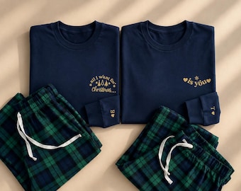 May include: Two navy blue long-sleeve shirts and two pairs of green and navy plaid pajama pants. The left shirt features gold text reading all I want for Christmas, and the right shirt reads is you. Each sleeve cuff has a gold letter and heart.