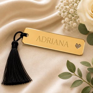 Könnte beinhalten: Ein rechteckiges, goldfarbenes Lesezeichen mit reflektierender Oberfläche, auf dem der Name ADRIANA in einer Serifenschrift eingraviert ist. Auf der rechten Seite befindet sich ein kleiner herzförmiger Ausschnitt und am linken Ende eine schwarze Quaste.