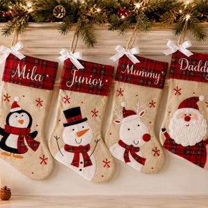 Può includere: Quattro calze natalizie beige con risvolti a quadri rossi e verdi, ciascuna con un fiocco di raso bianco e un nome ricamato in bianco. Le calze presentano personaggi applicati: un pinguino, un pupazzo di neve, una renna e Babbo Natale.